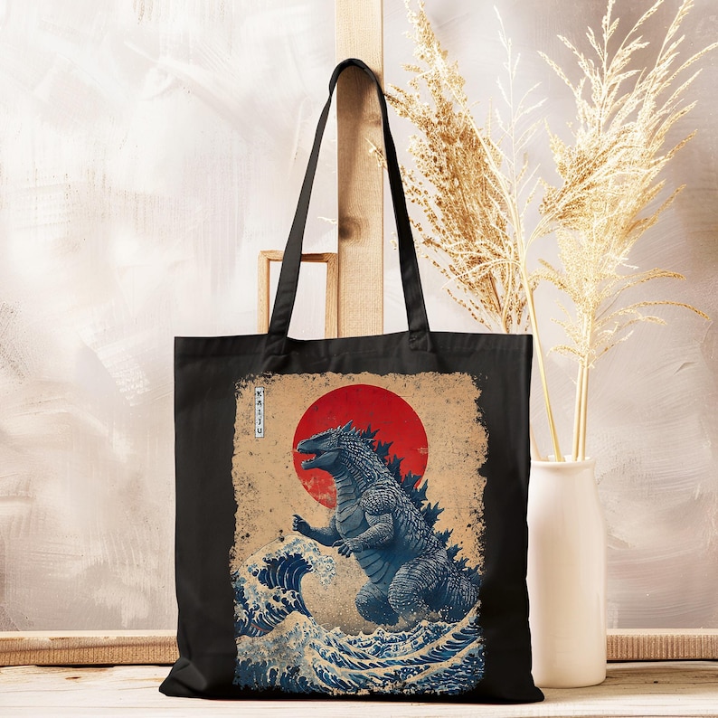 May include: Black tote bag with a blue Godzilla illustration, red sun, and ocean waves, inspired by Japanese art. The bag has long handles and a rectangular shape. The design includes Japanese text.