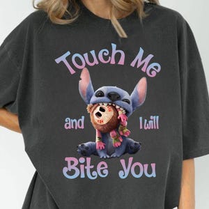 May include: Dark gray t-shirt featuring a cartoon image of Stitch biting a brown stuffed animal. The text "Touch Me and I Will Bite You" is printed above and below the image in a gradient font.