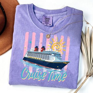 May include: A lilac Comfort Colors t-shirt with a cruise ship graphic and the words "Cruise Time". The design includes a sun, pink stripes, and blue stars. A gold necklace is draped across the shirt.