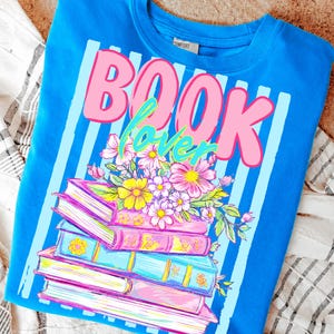 May include: A bright blue t-shirt featuring a graphic print of a stack of four books topped with a colorful floral arrangement. The text "BOOK lover" is printed in large pink and teal letters above the books, set against a light blue striped background.