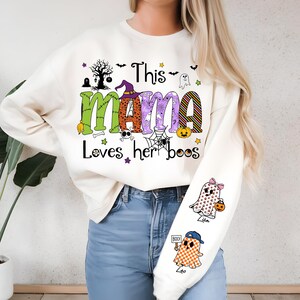 May include: Cream-colored Halloween-themed sweatshirt with the text "This MAMA Loves her boos" in green, orange, and purple. The design includes ghosts, a witch hat, a spider web, and pumpkins. The sleeve features two ghost graphics.