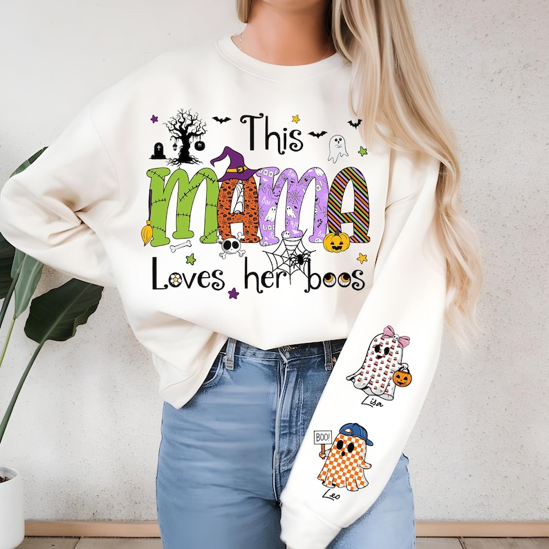 May include: Cream-colored Halloween-themed sweatshirt with the text "This MAMA Loves her boos" in green, orange, and purple. The design includes ghosts, a witch hat, a spider web, and pumpkins. The sleeve features two ghost graphics.