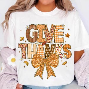 Może przedstawiać: Biała koszulka z napisem "GIVE THANKS" w jesiennym wzornictwie. Litery mają różne wzory, w tym kratę, kwiaty i panterkę, z dużą pomarańczową kokardą na dole. Jesienne liście i kwiaty są rozrzucone wokół tekstu.