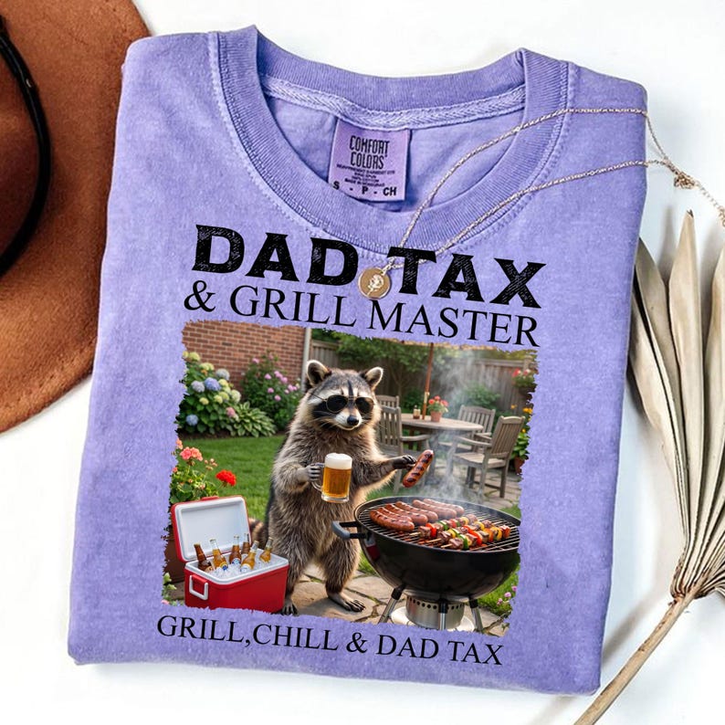 May include: A lilac Comfort Colors t-shirt with the text "DAD TAX & GRILL MASTER" and a raccoon graphic grilling. The raccoon wears sunglasses and holds a beer and a sausage. A red cooler with drinks is visible.
