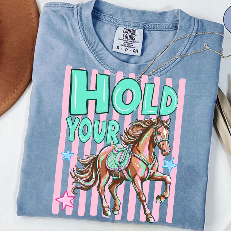 May include: A light blue Comfort Colors t-shirt with the words "HOLD YOUR" in turquoise and a brown horse graphic. Pink vertical stripes and star graphics are also present. A gold necklace is draped across the shirt.
