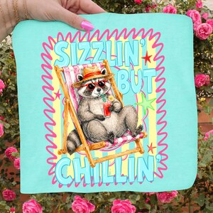 May include: A light blue beach towel with a raccoon illustration. The raccoon is wearing sunglasses and a hat, relaxing in a beach chair, holding a drink. The text "Sizzlin' But Chillin'" is displayed in a playful font.