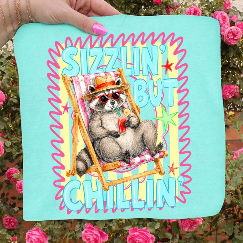 May include: A light blue beach towel with a raccoon illustration. The raccoon is wearing sunglasses and a hat, relaxing in a beach chair, holding a drink. The text "Sizzlin' But Chillin'" is displayed in a playful font.