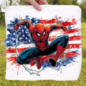 Puede incluir: Un tejido cuadrado blanco con una vibrante ilustración de Spider-Man. El superhéroe está representado en su clásico traje rojo y azul sobre un fondo de la bandera estadounidense, con salpicaduras de pintura roja, blanca y azul.