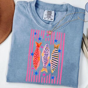 Pode incluir: Uma camiseta Comfort Colors azul clara com um design de peixes coloridos. O design inclui tr&ecirc;s peixes com listras verticais rosa e estrelas azuis. A camiseta tem gola redonda e mangas curtas. Um colar de ouro est&aacute; drapeado sobre a camiseta.