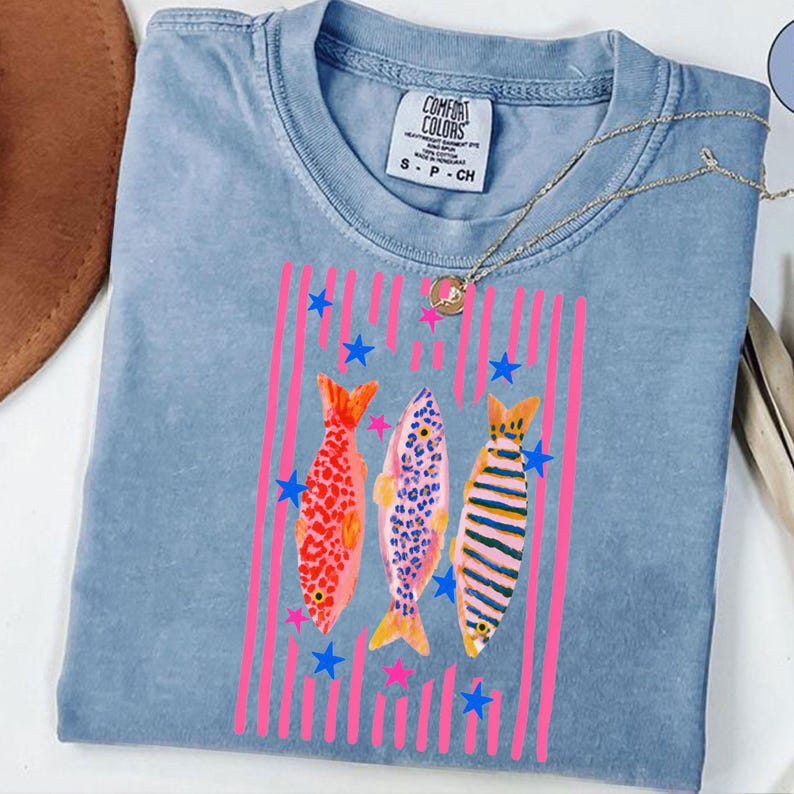 Pode incluir: Uma camiseta Comfort Colors azul clara com um design de peixes coloridos. O design inclui tr&ecirc;s peixes com listras verticais rosa e estrelas azuis. A camiseta tem gola redonda e mangas curtas. Um colar de ouro est&aacute; drapeado sobre a camiseta.