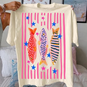 Pode incluir: Camiseta creme com um design de peixes coloridos. O gr&aacute;fico inclui tr&ecirc;s peixes em padr&otilde;es vermelhos, azuis e listrados, acentuados com listras verticais rosa e estrelas azuis. A camiseta &eacute; segurada por uma pessoa.
