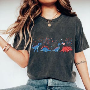 Puede incluir: Camiseta gris oscuro con una fila de dinosaurios de dibujos animados en azul y rojo, con fuegos artificiales y estrellas. Un dinosaurio sostiene una bandera americana. La camiseta es de estilo casual.