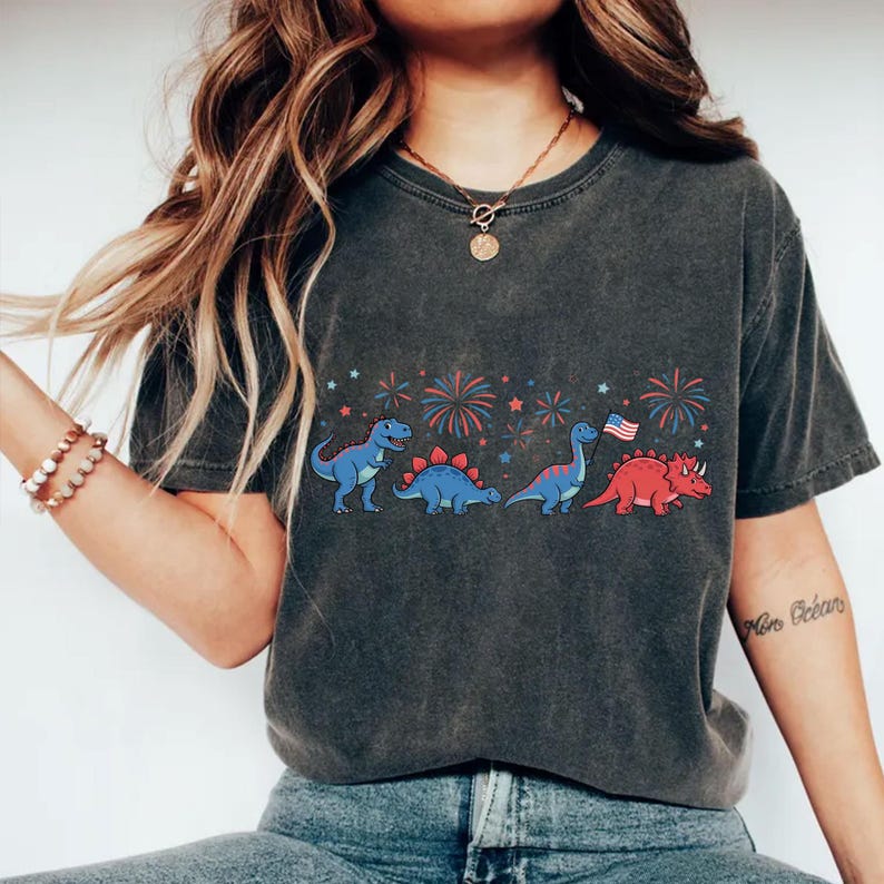Puede incluir: Camiseta gris oscuro con una fila de dinosaurios de dibujos animados en azul y rojo, con fuegos artificiales y estrellas. Un dinosaurio sostiene una bandera americana. La camiseta es de estilo casual.