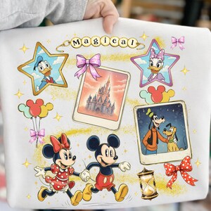 May include: White sweatshirt featuring a colorful Disney-themed design. The design includes Mickey and Minnie Mouse, Donald and Daisy Duck, Goofy and Pluto, and a castle. The word "Magical" is written across the top.