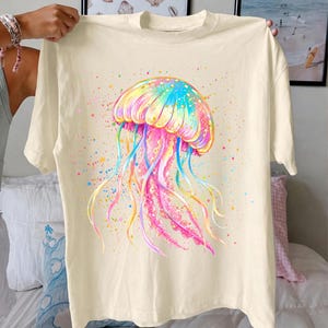 May include: Cream-colored t-shirt featuring a vibrant watercolor jellyfish design. The jellyfish showcases a colorful array of hues, including pink, blue, yellow, and green, with long, flowing tentacles. The design is accented with colorful paint splatters.