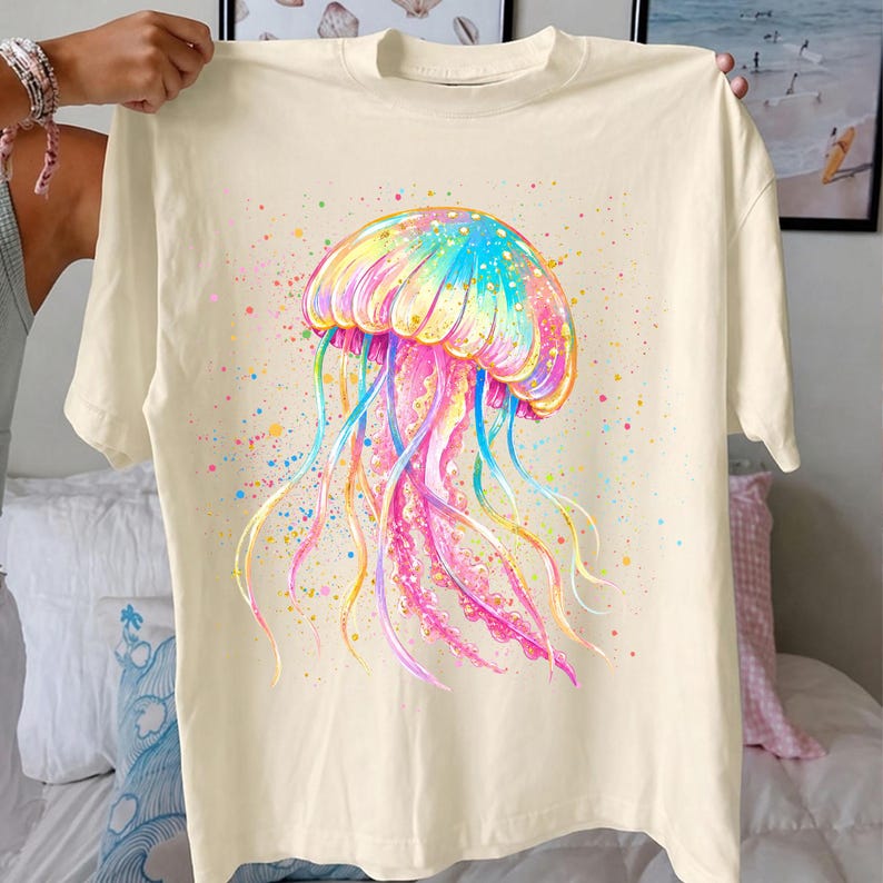 May include: Cream-colored t-shirt featuring a vibrant watercolor jellyfish design. The jellyfish showcases a colorful array of hues, including pink, blue, yellow, and green, with long, flowing tentacles. The design is accented with colorful paint splatters.
