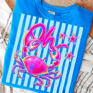 May include: A blue crew neck sweatshirt featuring a vibrant crab graphic with the word "Oh" in pink. The design includes pink stars and light blue vertical stripes, creating a playful, summery vibe.