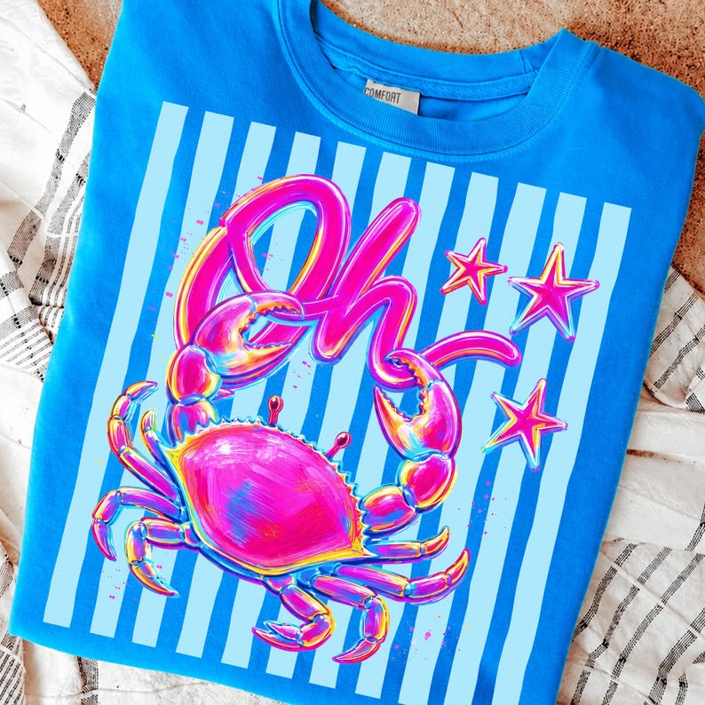 May include: A blue crew neck sweatshirt featuring a vibrant crab graphic with the word "Oh" in pink. The design includes pink stars and light blue vertical stripes, creating a playful, summery vibe.