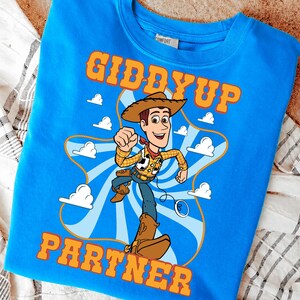 Puede incluir: Sudadera azul con un dise&ntilde;o de vaquero de dibujos animados. El dise&ntilde;o incluye las palabras "GIDDYUP PARTNER" en naranja. El vaquero lleva sombrero, chaleco y botas, sobre un fondo de nubes y una cuerda.