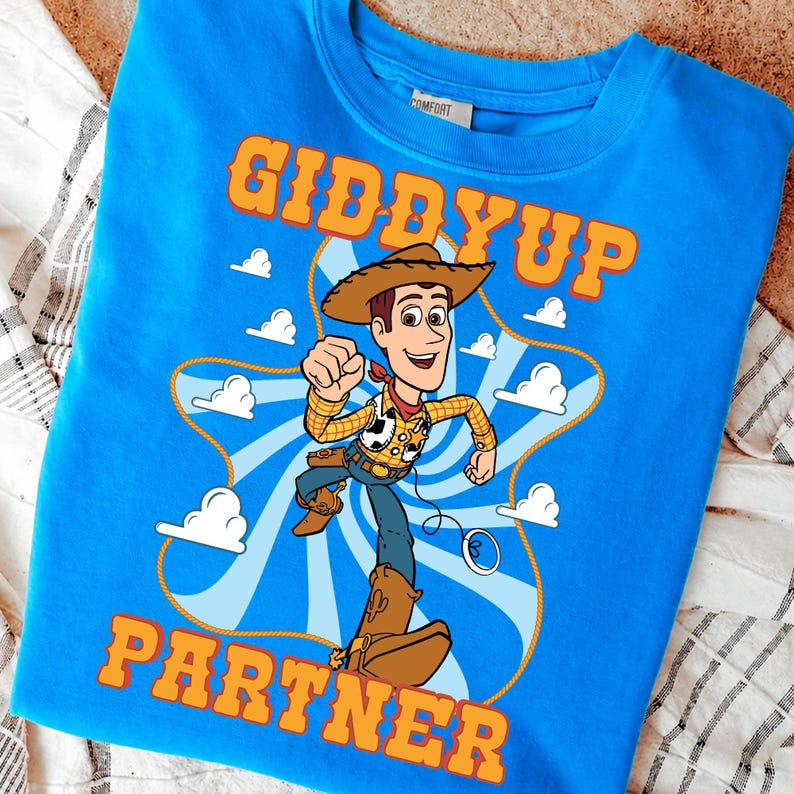 Puede incluir: Sudadera azul con un dise&ntilde;o de vaquero de dibujos animados. El dise&ntilde;o incluye las palabras "GIDDYUP PARTNER" en naranja. El vaquero lleva sombrero, chaleco y botas, sobre un fondo de nubes y una cuerda.