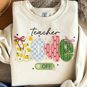 Op de afbeelding: Cr&egrave;mekleurige sweatshirt met de woorden "Teacher" en "WOE OFF" in een speels, kleurrijk ontwerp. De letters zijn versierd met citroenen, ruitpatronen, aardbeien en watermeloen. Geaccentueerd met strikken, bloemen en sterren.