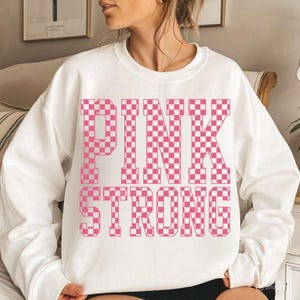 May include: White sweatshirt with the words "PINK STRONG" in a pink and white checkered pattern. The sweatshirt is a crew neck style and appears to be made of a soft material. The design is centered on the front of the shirt.