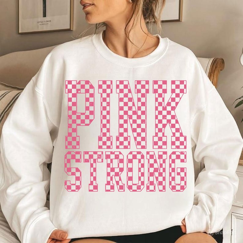 May include: White sweatshirt with the words "PINK STRONG" in a pink and white checkered pattern. The sweatshirt is a crew neck style and appears to be made of a soft material. The design is centered on the front of the shirt.