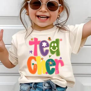 May include: A cream-colored t-shirt with the word "Teacher" in colorful, playful lettering. The design includes a smiley face, stars, and a butterfly. The shirt is worn by a child wearing sunglasses.
