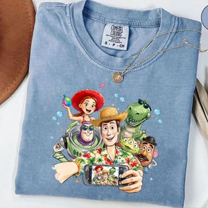 May include: Light blue Comfort Colors t-shirt featuring a graphic of characters from the Toy Story movie taking a selfie. The characters include Woody, Buzz Lightyear, Jessie, Rex, Mr. Potato Head, and the aliens. A gold necklace is draped across the shirt.