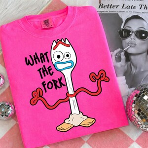 Puede incluir: Una sudadera rosa brillante con la frase "WHAT THE FORK" y un personaje de tenedor de dibujos animados. El personaje tiene un cuerpo blanco, detalles azules y rojos, y est&aacute; de pie sobre bloques de madera. La sudadera tiene una etiqueta Comfort Colors.