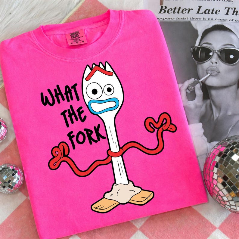 Puede incluir: Una sudadera rosa brillante con la frase "WHAT THE FORK" y un personaje de tenedor de dibujos animados. El personaje tiene un cuerpo blanco, detalles azules y rojos, y est&aacute; de pie sobre bloques de madera. La sudadera tiene una etiqueta Comfort Colors.