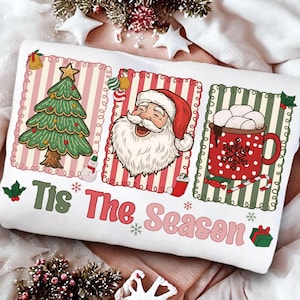 Puede incluir: Coj&iacute;n blanco con tem&aacute;tica navide&ntilde;a con tres ilustraciones enmarcadas. Los marcos presentan un &aacute;rbol de Navidad, Pap&aacute; Noel y una taza de chocolate caliente. El texto "Tis The Season" est&aacute; impreso en verde y rosa.