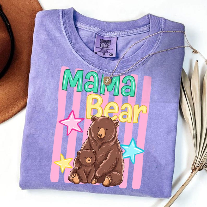 Pu&ograve; includere: Una maglietta lilla con le parole "Mama Bear" in verde e giallo, con un'illustrazione di un orso bruno e il suo cucciolo. Strisce rosa e bianche e stelle completano il design. La maglietta ha un girocollo e maniche corte.