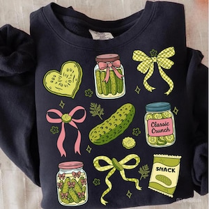 May include: Navy blue sweatshirt featuring a variety of pickle-themed illustrations. The design includes jars of pickles, a pickle heart, a pickle snack bag, and decorative bows. The illustrations are in shades of green, pink, and yellow.