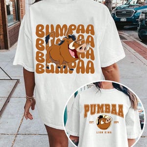 May include: White t-shirt with a cartoon image of Pumbaa from The Lion King. The shirt has the text "BUMPAA" repeated in brown letters. The second shirt is white with the text "PUMBAA" in brown letters and the text "EST. 1994 LION KING" in smaller brown letters.