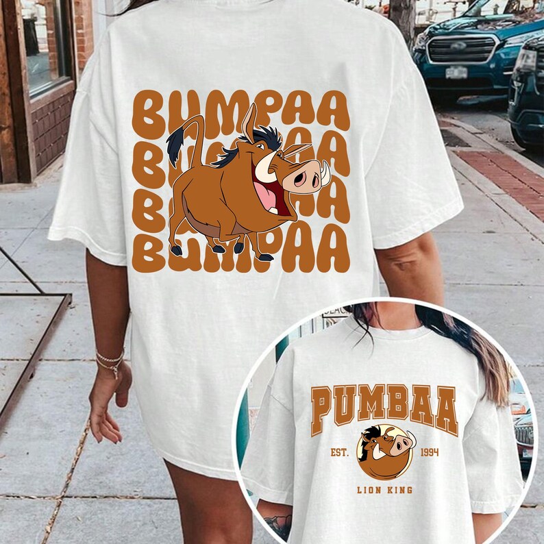 May include: White t-shirt with a cartoon image of Pumbaa from The Lion King. The shirt has the text "BUMPAA" repeated in brown letters. The second shirt is white with the text "PUMBAA" in brown letters and the text "EST. 1994 LION KING" in smaller brown letters.