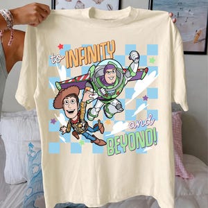 Puede incluir: Camiseta color crema con un gr&aacute;fico de Buzz Lightyear y Woody de Toy Story, con el texto "to INFINITY and BEYOND!". El dise&ntilde;o incluye estrellas y un fondo a cuadros.