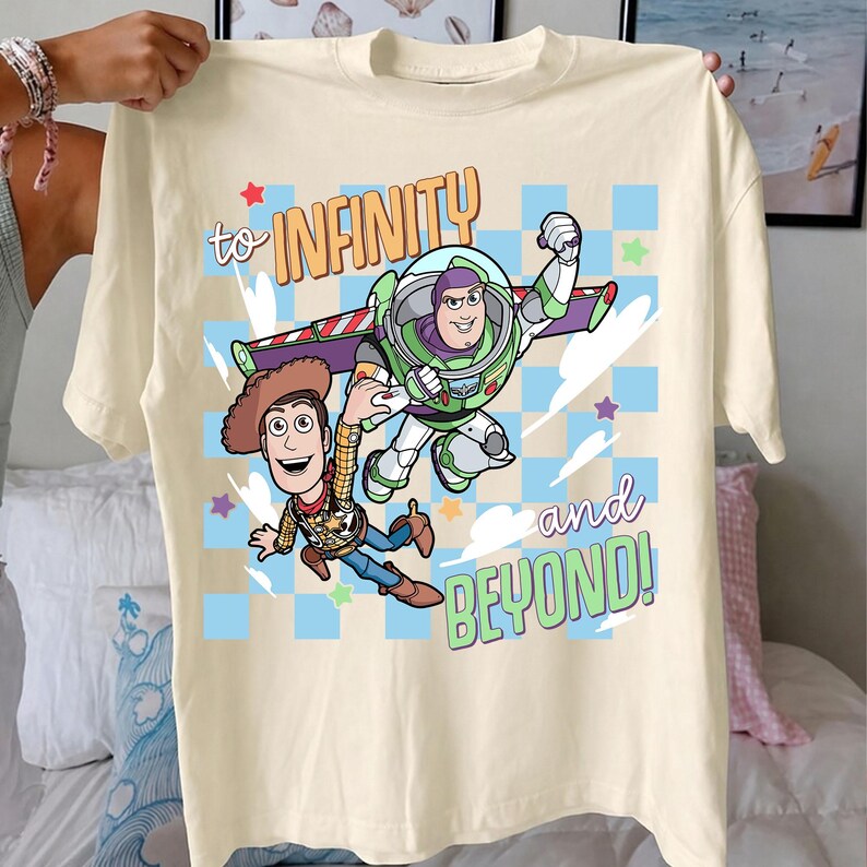 Puede incluir: Camiseta color crema con un gr&aacute;fico de Buzz Lightyear y Woody de Toy Story, con el texto "to INFINITY and BEYOND!". El dise&ntilde;o incluye estrellas y un fondo a cuadros.