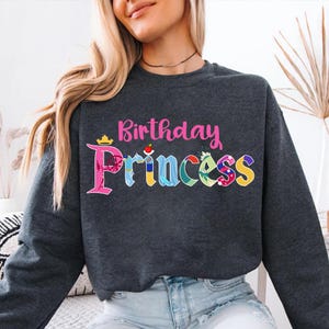 May include: A dark gray sweatshirt with the text "Birthday Princess" in pink lettering. The word "Princess" is made up of colorful images of Disney princesses.