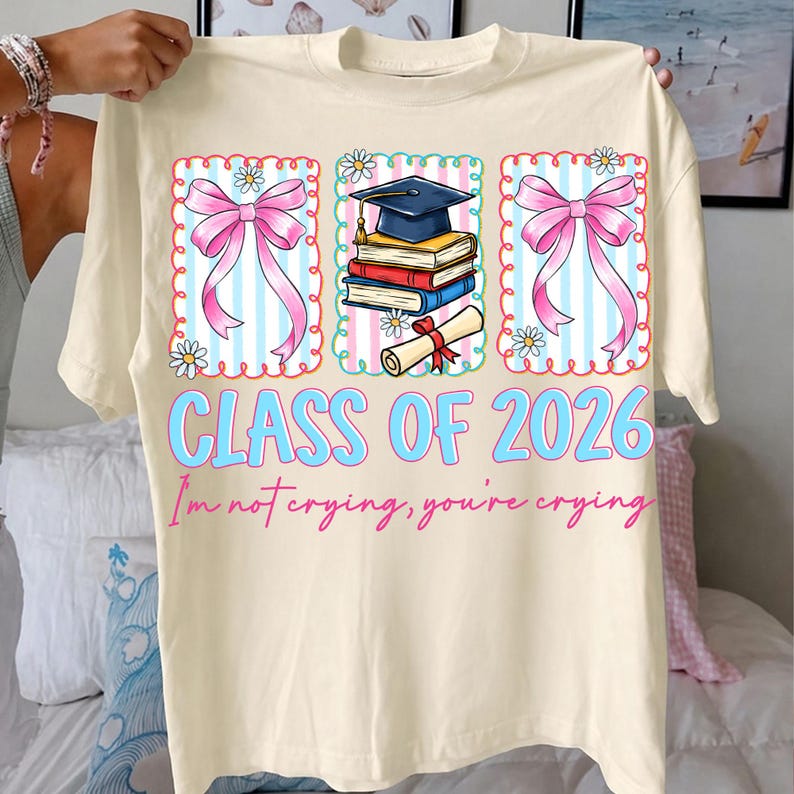 May include: Cream-colored t-shirt with a graduation theme. The shirt features three rectangular designs with pink bows, daisies, and a graduation cap, books, and diploma. The text "CLASS OF 2026" and "I'm not crying, you're crying" are in blue and pink.