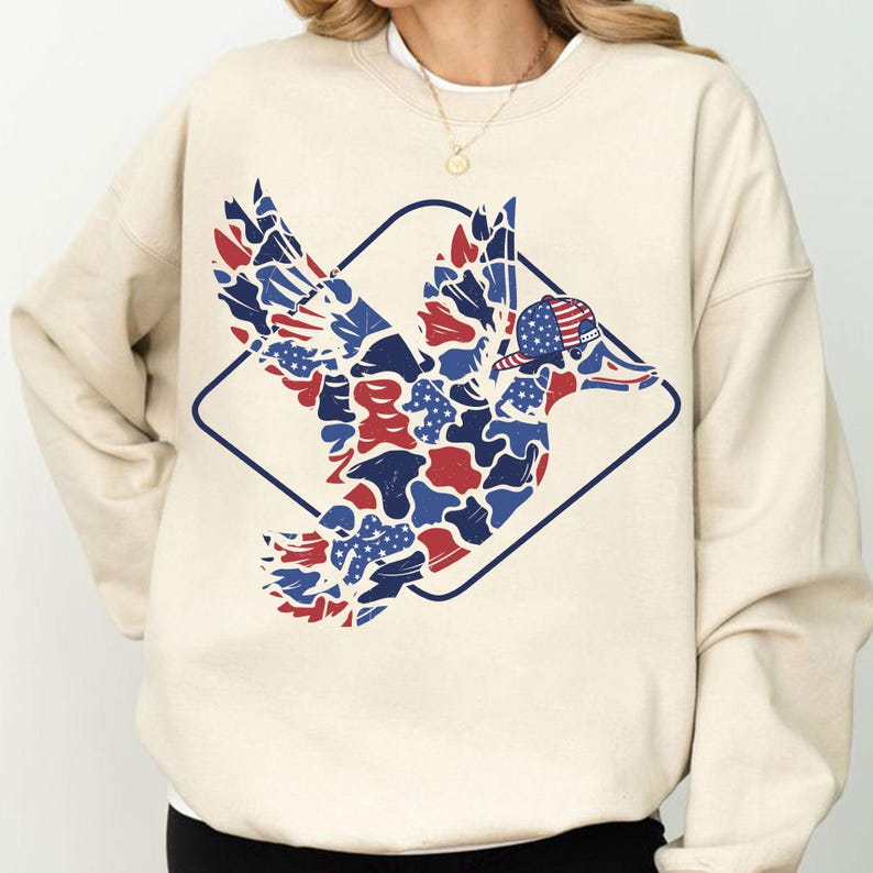 May include: Cream-colored sweatshirt featuring a patriotic duck design. The duck is composed of red, white, and blue shapes, with a matching hat. The design is framed by a dark blue diamond shape. The sweatshirt is a casual, comfortable style.