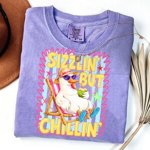 May include: A light purple Comfort Colors t-shirt featuring a cartoon duck wearing sunglasses and a hat, relaxing in a beach chair while holding a coconut drink. The shirt has the words "Sizzlin' But Chillin'" in yellow.