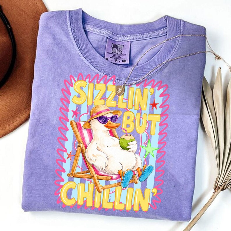 May include: A light purple Comfort Colors t-shirt featuring a cartoon duck wearing sunglasses and a hat, relaxing in a beach chair while holding a coconut drink. The shirt has the words "Sizzlin' But Chillin'" in yellow.