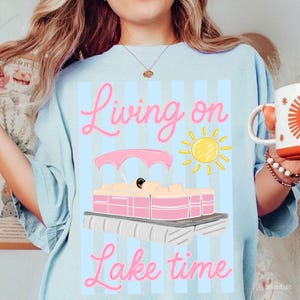 May include: Light blue sweatshirt with a pink and yellow graphic of a pontoon boat, sun, and the words "Living on Lake time" in pink script. The shirt has vertical blue stripes.