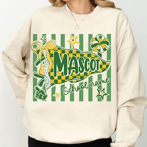 May include: A cream-colored crewneck sweatshirt featuring a graphic print of a green and yellow checkered pennant with the text "MASCOT" and "School name". The design includes a yellow bow, a pom-pom, stars, and a foam finger in green and yellow.