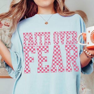 May include: Light blue t-shirt with the words "FAITH OVER FEAR" in large, pink checkered letters. The shirt is a soft, comfortable material. A white mug with an orange sun design is held in the hand.