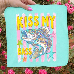 May include: A turquoise square textile with the text "KISS MY BASS" in yellow, with a colorful fish illustration. Pink and white stripes are behind the text and fish, with star accents in green, orange, and blue.