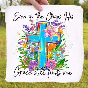 May include: White square fabric with a vibrant, turquoise cross design, surrounded by colorful flowers in pink, purple, orange, and blue. The text reads "Even in the Chaos His Grace still finds me."