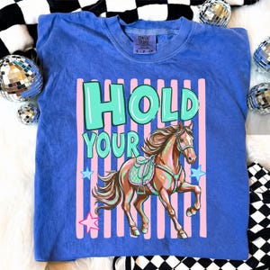 May include: Blue crew neck t-shirt with the words "HOLD YOUR" in turquoise and a brown horse graphic. The horse has a saddle and is set against a pink and white striped background with blue stars.