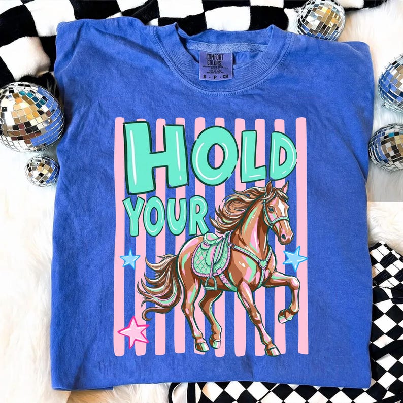 May include: Blue crew neck t-shirt with the words "HOLD YOUR" in turquoise and a brown horse graphic. The horse has a saddle and is set against a pink and white striped background with blue stars.